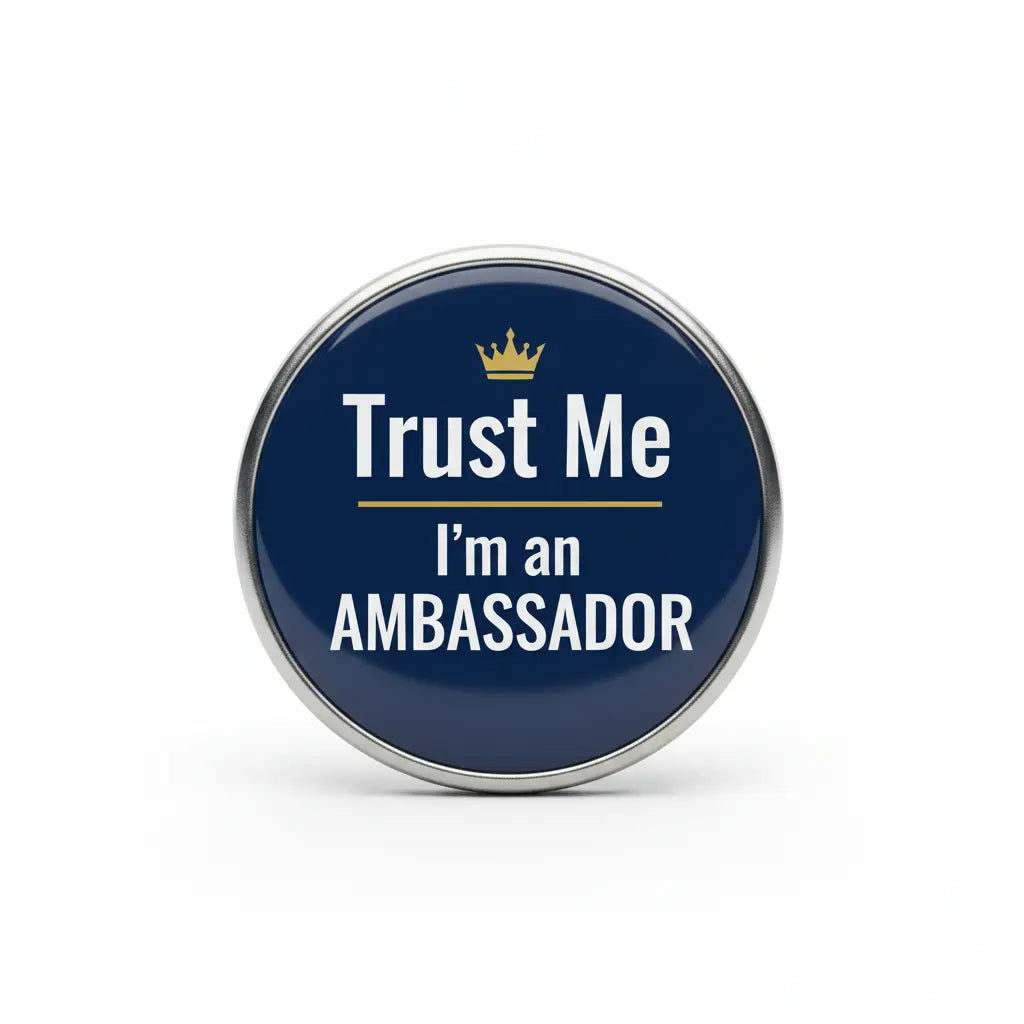 Round pinback button badge with dark blue face, silver rim, “Trust Me I’m an AMBASSADOR” text and gold crown