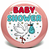 A round pink baby shower button badge featuring a white stork carrying a blue baby bootie and the words 'BABY SHOWER' in red and blue lettering.