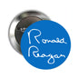 Round gold-tone pinback badge with Ronald Reagan signature in white on blue disc