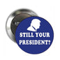 Blue and silver pinback button with white text "STILL YOUR PRESIDENT?"