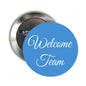 Round pinback button with blue face and cursive "Welcome Team" text, silver back visible