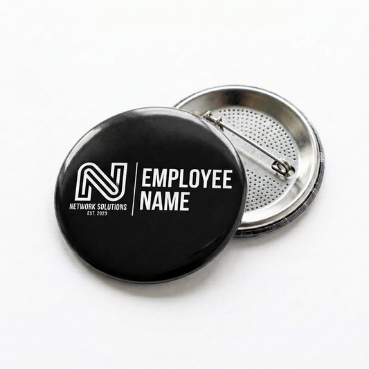 Two round pinback buttons on a white background. The front button is black with a white stylized 'N' logo and the text 'EMPLOYEE NAME' and 'NETWORK SOLUTIONS EST.