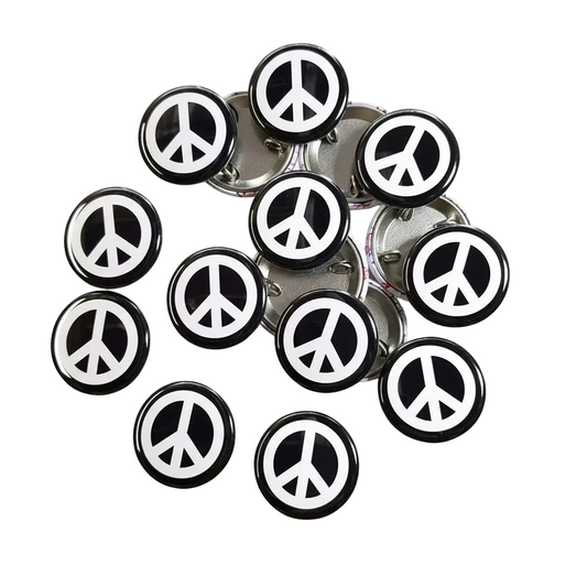 A group of round black and white peace sign lapel pins with silver metal backing, arranged on a checkered gray background with a blue border.