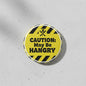 Round pinback button with yellow CAUTION: May Be HANGRY text and black hazard stripes