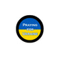 Round pinback button with black rim, blue/yellow center text "PRAYING FOR UKRAINE"