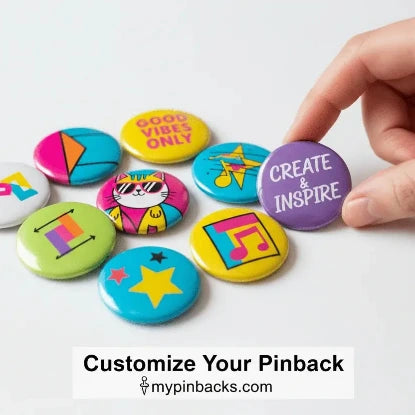 A hand is placing a purple pinback button with the text 'CREATE & INSPIRE' next to a cluster of other colorful pinback buttons on a white background. A banner at the bottom reads 'Customize Your Pinback' with 'mypinbacks.