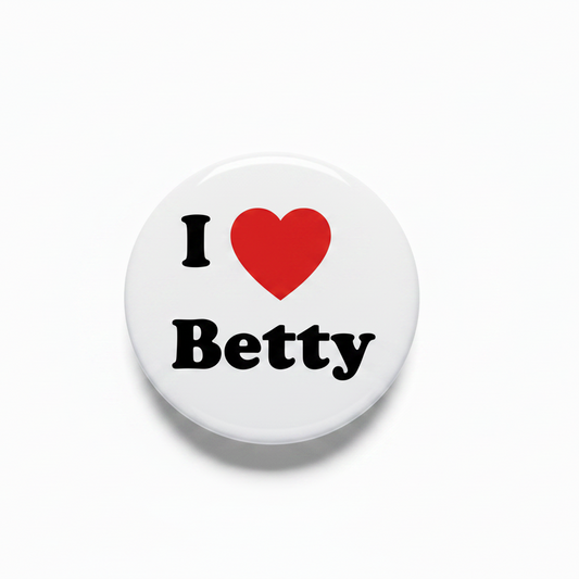 A round pin-back button with a silver metal rim and safety pin clasp, featuring a white face with the text 'I' in black, a red heart, and 'Betty' in black.