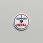 A round white button badge featuring the text 'I SUPPORT NEPAL' with a red and blue heart symbol in the center, shown on a light gray background.