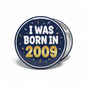 Round navy pinback button with white text “I WAS BORN IN 2009” and metallic edge