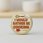 Round pinback button with chef utensils and 'I WOULD RATHER BE COOKING'.