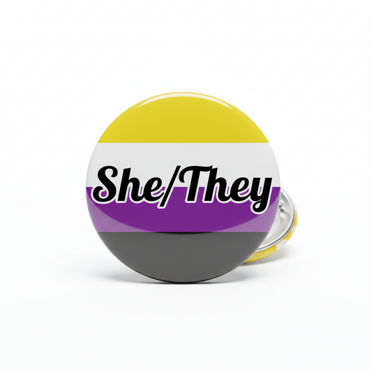 Rainbow circular pinback button with She/They lettering and metal pin back, black trim