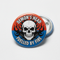 A round pin-back button badge featuring a white skull over red and blue flames, with the text 'DEMON'S HEAD' at the top and 'FUELLED BY FIRE' at the bottom. The front of the button is shown on a light gray background, with the back metal pin visible behind it.