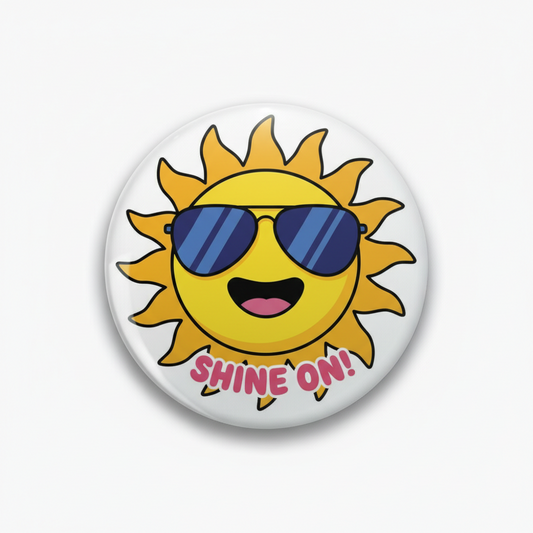 A round pin-back button badge featuring a cartoon sun with blue sunglasses and the text 'SHINE ON!' in pink, shown on a light gray background.