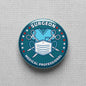 Circular teal surgeon medical lapel pin with mask, scrubs icons, and text