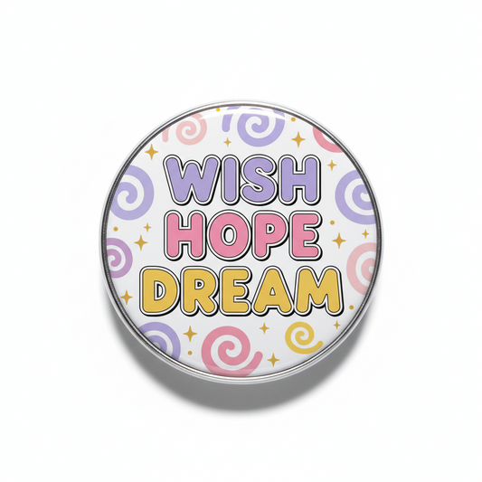 A round pinback button with a silver metal border on a white background. The button features the words 'WISH HOPE DREAM' in stylized, colorful lettering (purple, pink, and yellow) with a pattern of pastel swirls and small gold stars behind it.